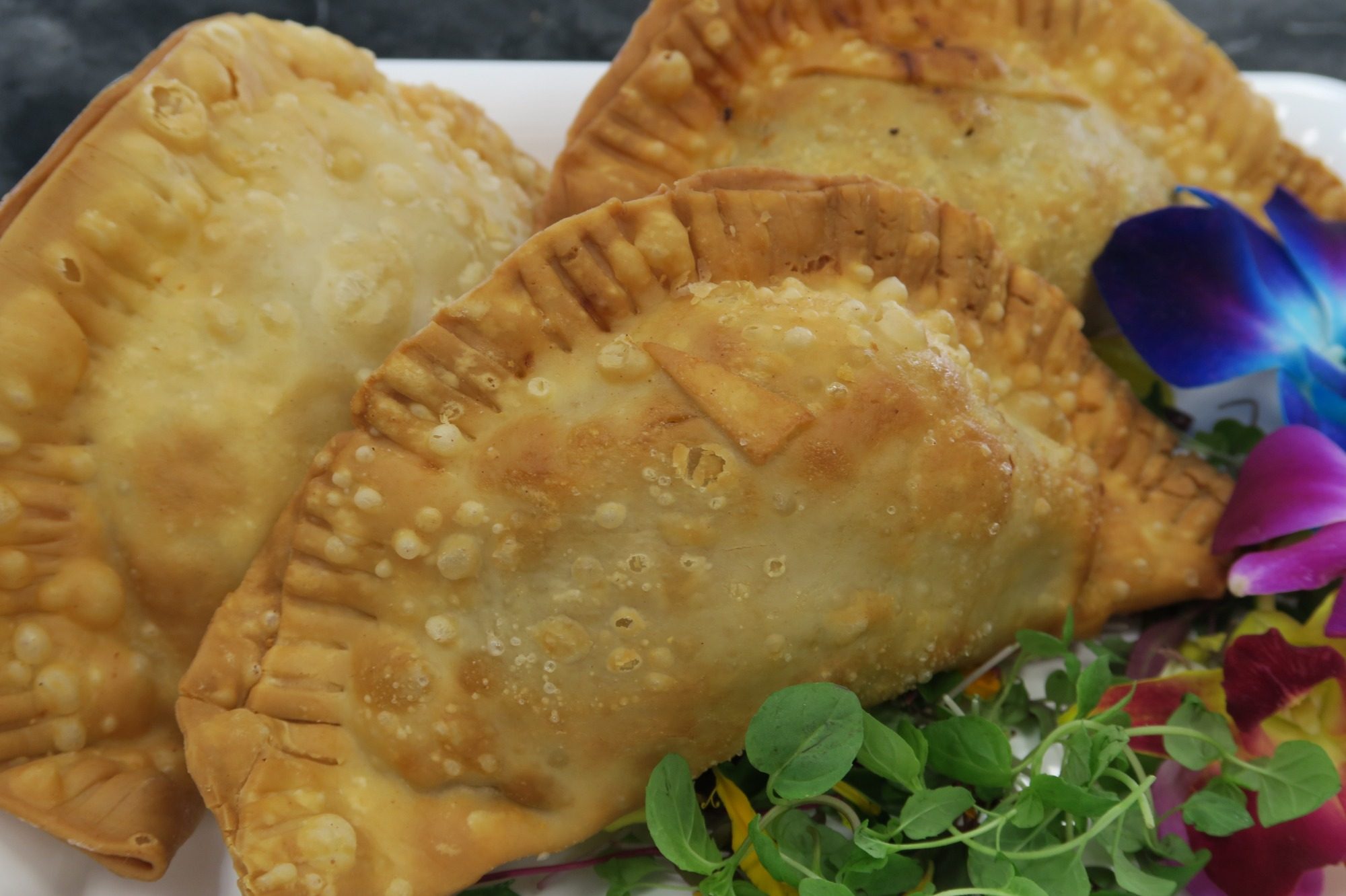 Empanadas Three golden-brown empanadas on a plate with colorful edible flowers and greens.